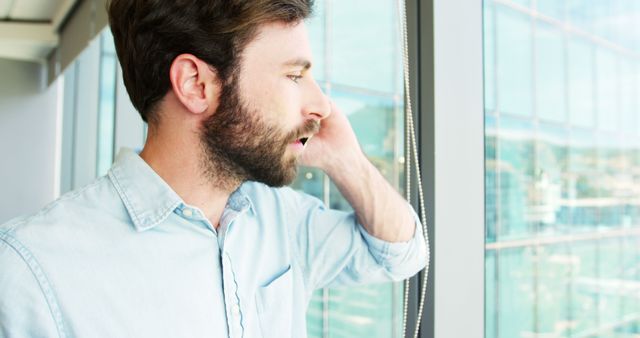 Young man standing by an office window looking outside with a contemplative expression. Beard and casual attire suggest a modern professional setting. Can be used for business, career, and productivity themes, as well as themes related to thoughts, ideas, and planning.