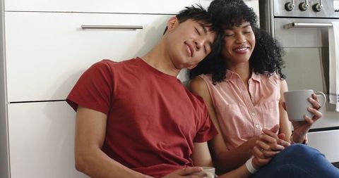 Couple leaning together on kitchen floor holding mugs and hands, cozy intimate morning