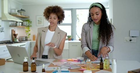 Women entrepreneurs collaborating on skincare packaging prototypes and brand design in kitchen works