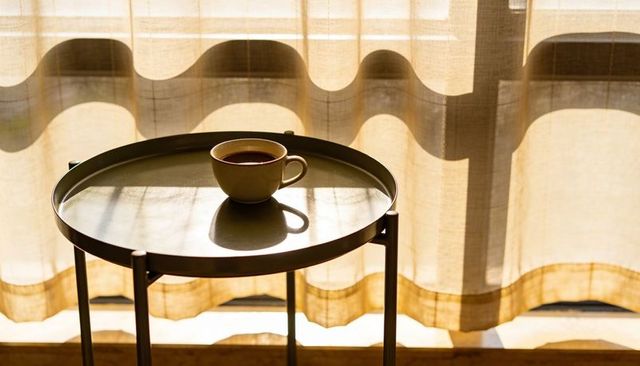 Morning coffee casting soft shadow on round tray table backlit by beige curtains