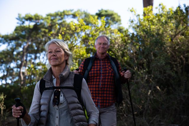 Senior couple enjoying outdoors, hiking with Nordic walking sticks in a lush forest setting. Perfect for promoting senior health, active aging, fitness activities, and recreational travel.