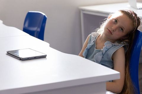 Young girl sitting at classroom desk daydreaming beside tablet in soft natural light