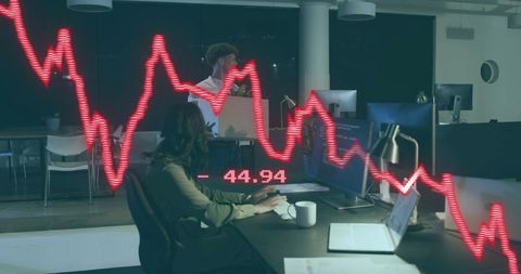 Late-night analyst typing at desk while colleague carrying box with plunging red stock graph