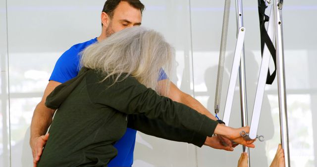 Senior woman exercising with assistance from a personal trainer in a Pilates studio. Ideal for promoting fitness and wellness among older adults, personal training services, and rehabilitation programs.