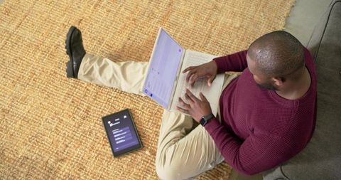 African American man typing on laptop sitting on rug with tablet nearby for remote work
