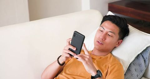 Asian man reclining on sofa using smartphone for messaging and streaming in living room
