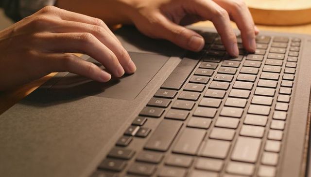 Typing on laptop keyboard hands using trackpad on wooden desk under warm light