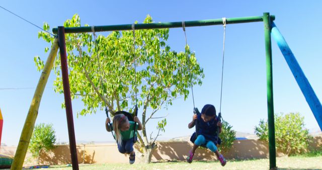 Two children are enjoying a sunny day at the playground, swinging high on a swing set, with copy space. Their joyful activity captures the essence of carefree childhood moments outdoors.