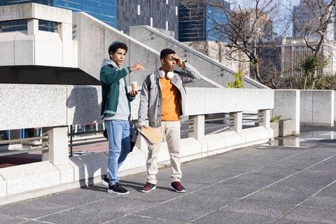 African american teen friends standing on urban plaza pointing, holding coffee and notebooks