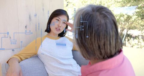 Korean woman talking, leaning on couch, touching temple with blue circuit overlay