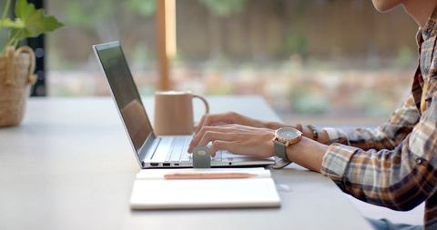 Remote worker typing on slim laptop at sunny home workspace with notebook and coffee