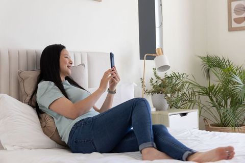 Mature asian woman reclining on bed smiling and holding smartphone in sunlit minimalist bedroom