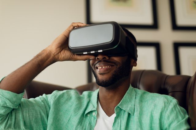 African-American man sitting on a leather chair indoors, wearing VR goggles and smiling. Ideal for use in articles or advertisements related to virtual reality technology, modern entertainment, or digital innovation. Can also be used in lifestyle blogs or tech reviews.