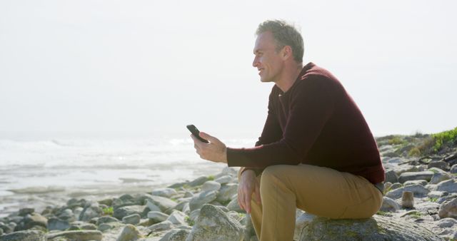 Happy senior caucasian man sitting on sunny rocky beach using smartphone, copy space. Communication, retirement, vacations, wellbeing and active senior lifestyle, unaltered.