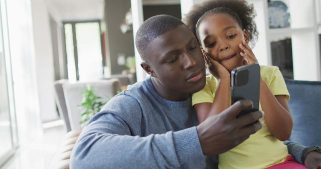 Father engaging with his daughter while sharing moments on a smartphone. Ideal for use in family-related content, technology discussion, parenting articles, and lifestyle blogs focusing on bonding and modern communication.