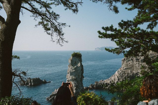 Photo shows tourists viewing a stunning coastal rock formation and the ocean. Rocky cliffs and green trees frame the scene, giving it a picturesque and serene feel. Ideal for travel magazines, tourism advertising, posters, and websites promoting nature trips and scenic destinations.