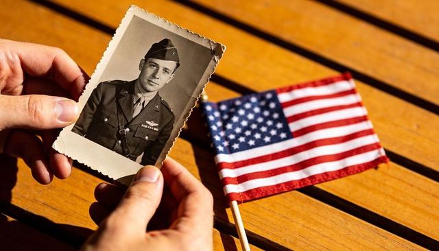 Hands holding vintage wwii portrait of serviceman beside small american flag on sunlit table