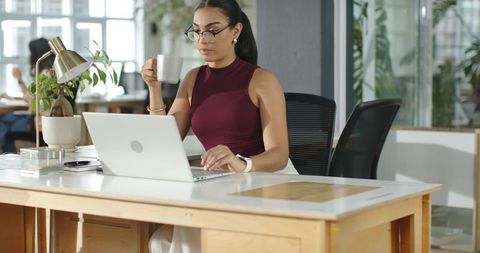 Young Professional Working on Laptop Holding Coffee Mug in Sunlit Coworking Space