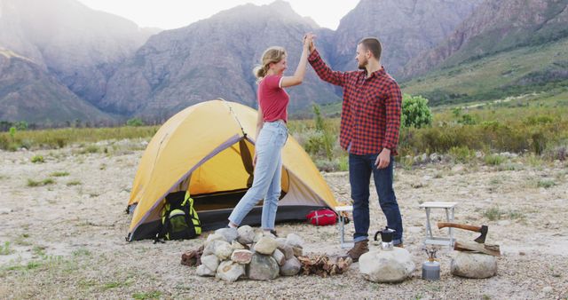 Young couple high-fiving in front of yellow tent in scenic mountain landscape. Camping gear and fire pit are nearby. Perfect for promoting outdoor adventure, camping vacations, and bonding experiences in nature.