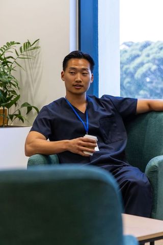 Healthcare worker relaxing in staff lounge wearing navy scrubs holding coffee cup and lanyard