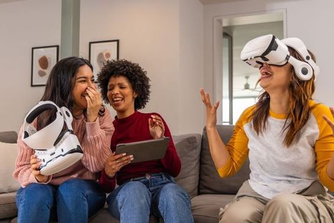 Diverse happy friends laughing while testing vr headsets together in cozy home living room