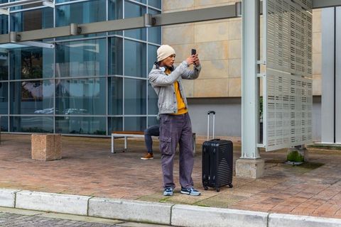 Urban traveler checking smartphone beside wheeled suitcase outside modern glass building