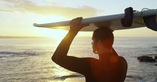 This visual captures a surfer holding his surfboard while watching a stunning sunset by the ocean shore. The golden hour light adds a warm and serene ambiance perfect for travel, adventure, lifestyle, and outdoor activity themes. Suitable for use in promotional material for beach vacations, surf courses, and motivational content centered around nature and outdoor sports.