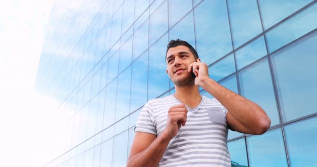 This stock photo features a confident young man talking on his smartphone in front of a modern glass building. Useful for business communication themes, urban lifestyle, technology usage, and corporate presentations.