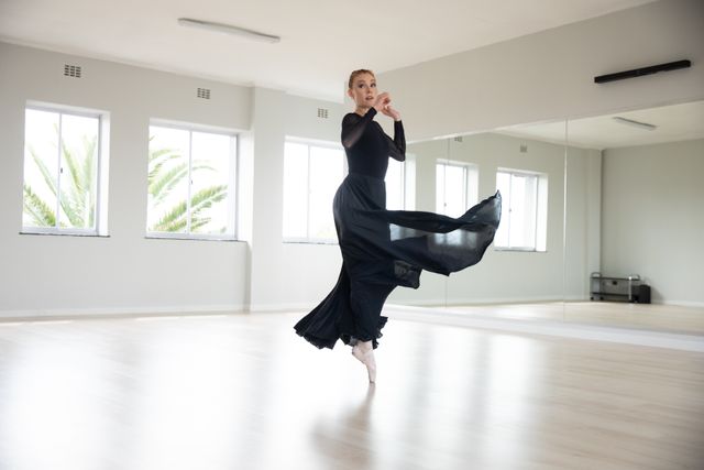 Caucasian female attractive ballet dancer warming up and practicing in a bright ballet studio, dancing black dress standing on her toes. Focused on her exercise, preparing for a class.