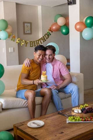 Mixed-race male couple celebrating birthday on sofa with mini cake and pastel balloons