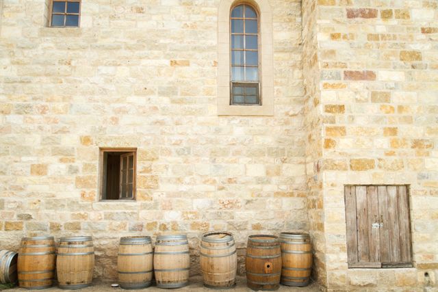 Old wooden barrels lined up next to rustic stone building with arched windows and a wooden door. This photo can be used for illustrating concepts related to wine making, vintage storage, historical architecture, or rustic outdoor settings.
