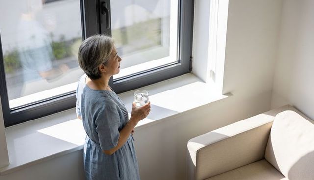 Mature woman standing at sunlit window holding glass of ice water, contemplative morning