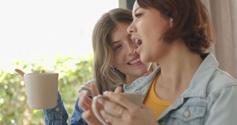 Smiling female friends holding coffee mugs by sunlit window, sharing warm morning moment