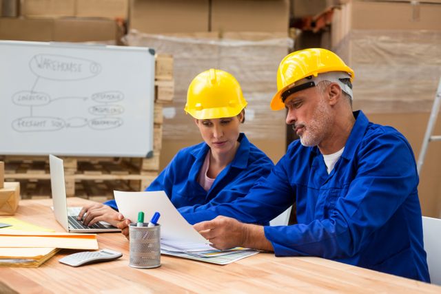 Warehouse workers in blue uniforms and hard hats are discussing documents at a table in a warehouse. They are engaged in planning and collaboration, with a whiteboard and stacks of boxes in the background. This image can be used to depict teamwork, logistics, and business management in an industrial setting.