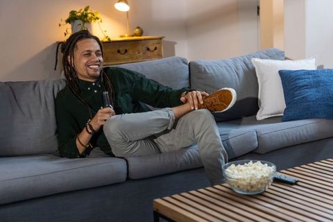 Relaxing african american man lounging on sofa with beer and popcorn in cozy living room