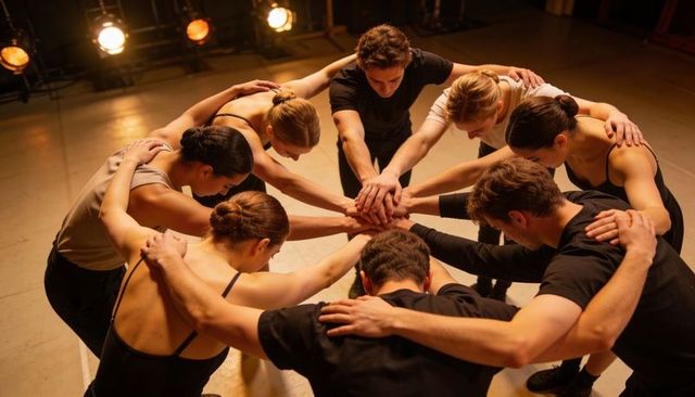 Dancers Huddling in Circle Stacking Hands on Marley Floor under Warm Stage Lights