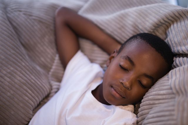 This image shows a young boy peacefully sleeping on a couch at home. He is wearing a white shirt and appears to be very comfortable. This image can be used for themes related to childhood, relaxation, home life, and peaceful moments. It is ideal for use in articles, blogs, and advertisements focusing on family life, sleep health, and child well-being.