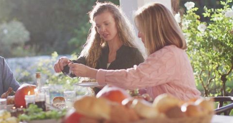 Two women passing salad dressing at outdoor garden lunch table with bread and candles
