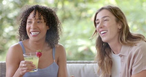Multiracial friends laughing on outdoor patio sofa sharing refreshing strawberry drink