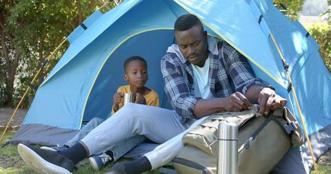 African american father and son camping together at blue tent sharing mug and packing duffel