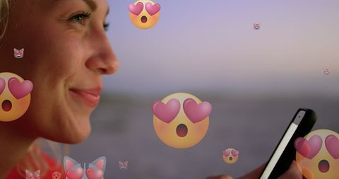 Joyful woman engaging with emojis on smartphone at sunset beach