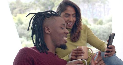 Couple sharing coffee and smartphone on couch, smiling and relaxing in cozy home