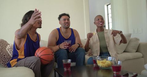 Friends watching basketball game on couch cheering, holding basketball, snacks and drinks