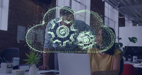 Team collaborating around holographic cloud gears while working on laptop in open office