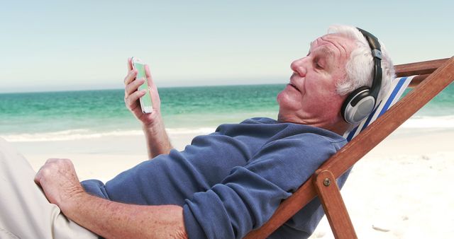 Senior man enjoying a relaxing day on the beach while listening to music with headphones. Ideal for themes related to retirement, leisure, travel, technology use among elderly, and summertime relaxation.