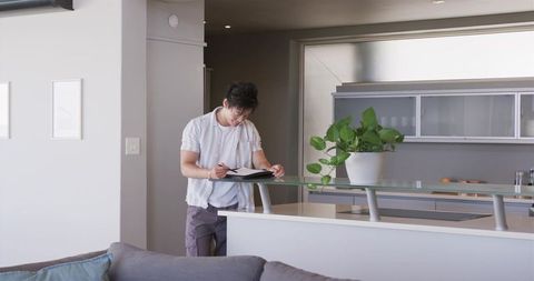 Asian man writing in notebook at modern minimalist kitchen island with glass railing