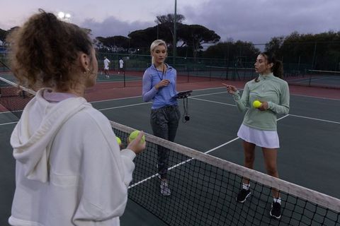 Women coaching tennis practice at dusk on outdoor hard court with clipboard and balls