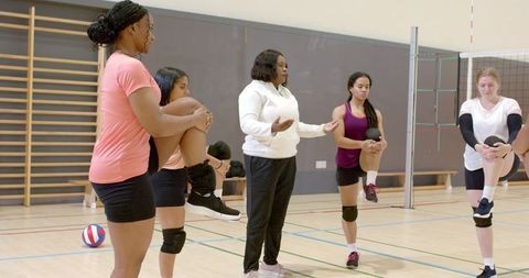Diverse female volleyball team stretching and balancing while coach gesturing in gym