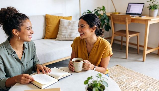 Two friends smiling and chatting over coffee in cozy living room workspace with notebook