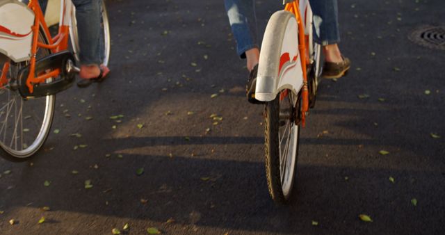 Focus on two people riding orange bicycles on a bike path. The close-up view features wheels and legs, with late afternoon sunlight casting long shadows. Perfect for topics related to outdoor activities, health and fitness, urban transportation, or recreational cycling.